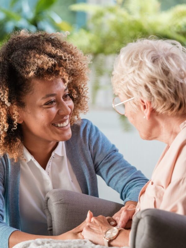 Smiling young female caregiver holding hands and talking with senior woman in living room